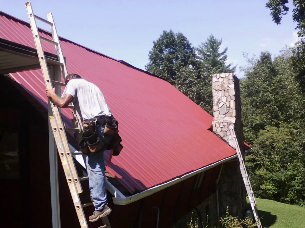 Roofer on a ladder beside a red metal roof and stone chimney — Soffit & Fascia Repair by Roofing Brothers in Ottawa Lake